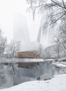 a pond surrounded by snow covered trees and tall buildings in the background with water reflecting it