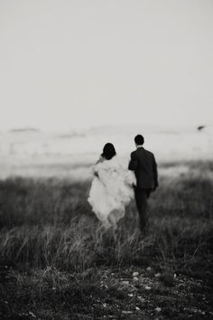 a man and woman walking through a field holding hands in black and white photo with the sky behind them