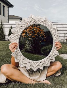 a woman sitting in the grass holding up a mirror to her face with an orange flower on it