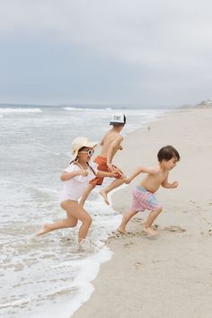 three children running on the beach with one child wearing a hat