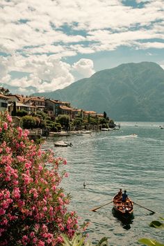 two people in a row boat on the water with mountains in the background and flowers in the foreground