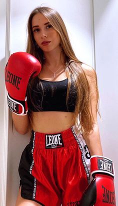 a beautiful young woman wearing red boxing shorts and black top standing next to a white wall