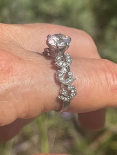 a woman's hand with a diamond ring on top of her finger, in front of some grass