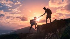 two people holding hands on top of a hill with the sun setting in the background