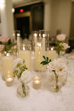 there are many candles and vases with flowers in them on the white table cloth