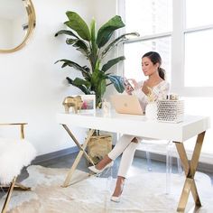 a woman sitting at a white desk with a laptop computer on her lap and potted plant in the corner