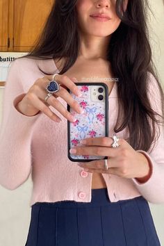 a woman wearing a pink sweater and blue skirt holding up her cell phone