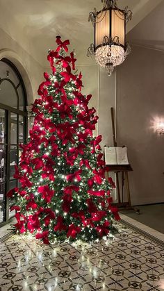 a decorated christmas tree with red bows and lights in a large room next to a chandelier