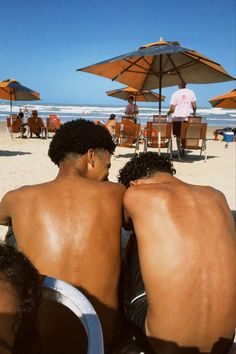 two men sitting on the beach under umbrellas looking out at the water and sand