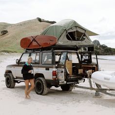 a woman standing next to a vehicle with a boat on the beach in front of it