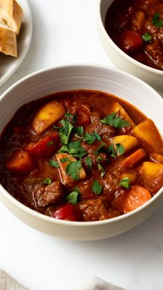 two bowls filled with stew and vegetables on top of a white table next to bread