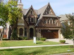 a large brick and stone house with two garages on the front lawn, surrounded by trees