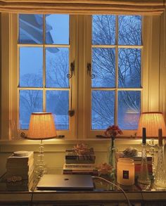 a desk with two lamps and books on it in front of a window that has snow outside