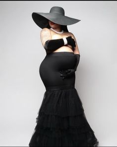 a woman in a black dress and hat with her hands on her hips, standing against a white background