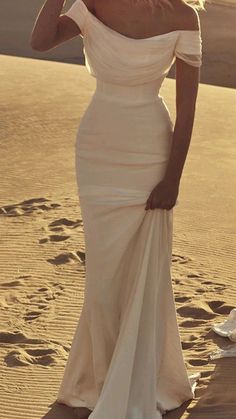 a woman standing in the sand wearing a white dress with off the shoulder and long sleeves