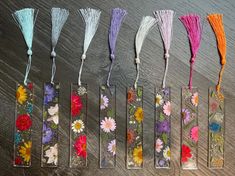 six flower bookmarks with tassels in them on a wooden surface, lined up next to each other