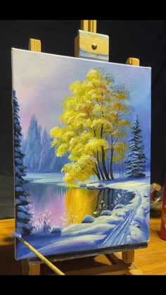 an easel with a painting on it that has a yellow tree in the snow