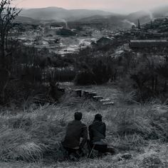 two people sitting in the grass looking at an industrial area