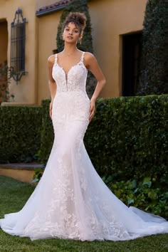 a woman in a white wedding dress standing on the grass near a house with bushes