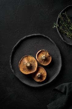 three mushrooms on a black plate next to a bowl with sprigs of herbs