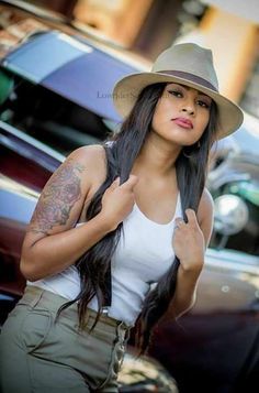 a woman with long hair wearing a hat and posing for the camera in front of parked cars