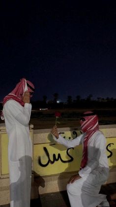 two men dressed in white and red sitting on a bench with one holding a flower