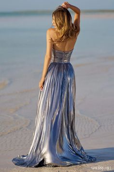 a woman in a long silver dress standing on the beach looking at the water with her back to the camera