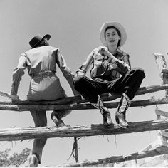 two people sitting on a wooden fence with hats and boots around their necks, one wearing a cowboy hat