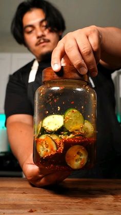 a man holding a jar filled with sliced cucumbers on top of a wooden table