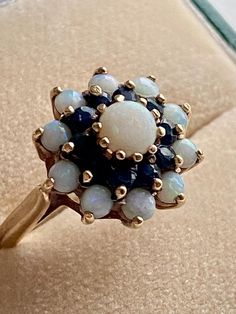 A beautiful 9CT Gold ring with an unusual combination of sapphires and white opals to create this unique floral cluster ring and the contrasting gemstones look perfect together. The stones are on a raised cage and the band is plain.  The top measures 1/2 " diameter or 1.2 cm. U.K Size Q, U.S size 8 1/4 FULL ENGLISH hallmarks. 1980s. There is a chip to one of the sapphires which can only be seen using a loop. Otherwise the ring is in GOOD vintage condition. We will wrap your order with care. We o Vintage Multi-stone Opal Ring Gift, White Cluster Ring Stamped 14k, Vintage Multi-stone Cluster Ring, Vintage Gemstone Cluster Ring, Antique Multi-stone Cluster Jewelry, Vintage White Cluster Ring With Gemstone, Vintage Multi-stone Opal Rings, Vintage Opal Multi-stone Rings, Vintage White Opal Ring Collectible