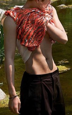 a man with no shirt standing in front of a body of water wearing an orange sequin top