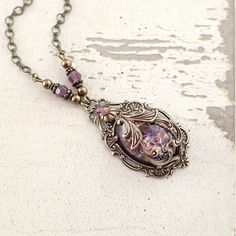 "To make this neo Victorian style necklace, I wrapped an amethyst opal glass cabochon in exquisitely detailed antiqued brass filigree. The stone is set in an antique Victorian style setting, which I embellished with matching opalescent Swarovski crystal stones. I finished the necklace with Swarovski beads, pearls, and vintage style infinity-link chain. --------------------------------------------------------------------------------------------- Dimensions and details - Focal cabochon measures 18 Victorian Brass Cabochon Jewelry, Vintage Brass Necklaces With Cabochon, Antique Gold Cabochon Necklace, Antique Gold Necklace With Cabochon, Antique Bronze Necklace With Cabochon, Handmade Vintage Amethyst Jewelry, Antique Purple Metal Jewelry, Antique Handmade Purple Jewelry, Handmade Antique Purple Jewelry