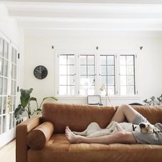 a man laying on top of a brown couch next to a white wall with windows