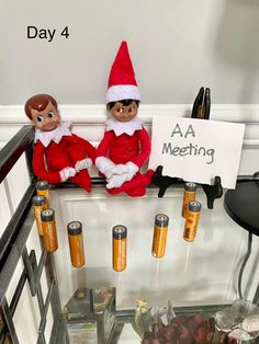 two elfs sitting on top of a shelf with batteries in front of them and a sign that says aa meeting