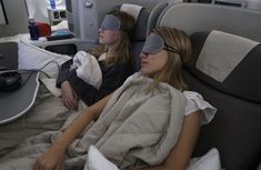 two women with blindfolds are laying on an airplane bed and one is wearing a sleeping mask