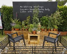 an outdoor garden with potted plants and two chairs in front of a sign that says,