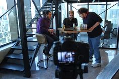 two men sitting at a table in front of a camera and another man standing next to them