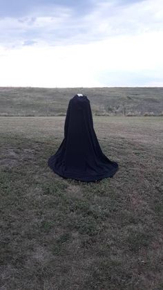 a person in a black cloak sitting on top of a grass covered field with a sky background