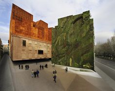 people are walking around in front of a building with a large green wall on it's side