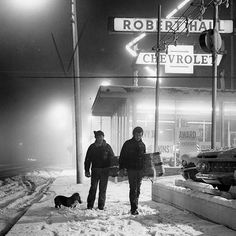 two people standing in the snow next to a car and street sign at night time