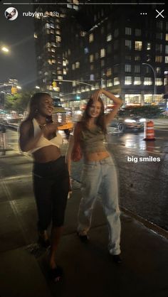 two women are standing on the sidewalk at night, one is holding her hand up to her head