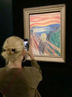 a woman taking a picture of a painting with her cell phone while standing in front of it