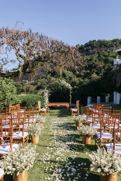 an outdoor ceremony set up with wooden chairs and white flowers on the aisle, surrounded by greenery