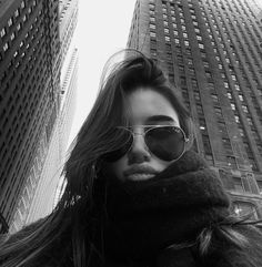 a woman wearing sunglasses standing in front of two tall buildings with her hair blowing in the wind