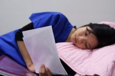 a woman is laying in bed reading a piece of paper with her head on the pillow