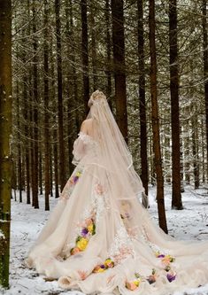 a woman in a wedding dress is standing in the snow near some trees and flowers