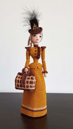 a crocheted doll holding a purse and wearing a hat with feathers on it