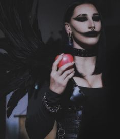 a woman with black makeup holding an apple in her right hand and wings on her left shoulder