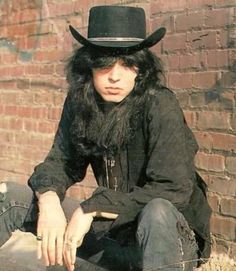 a man with long hair wearing a black hat and sitting on the ground next to a brick wall