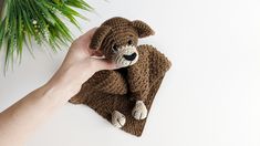 a hand holding a crocheted teddy bear on top of a brown blanket next to a green plant